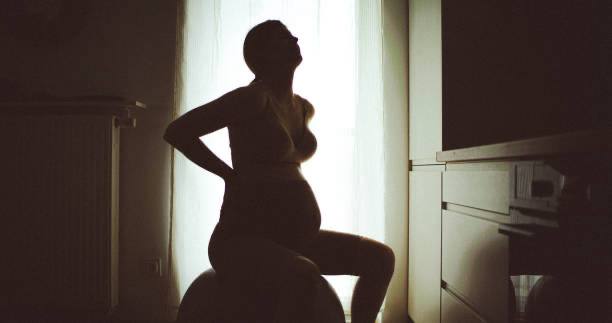 Pregnant woman silhouetted against a window, seated on a birth ball