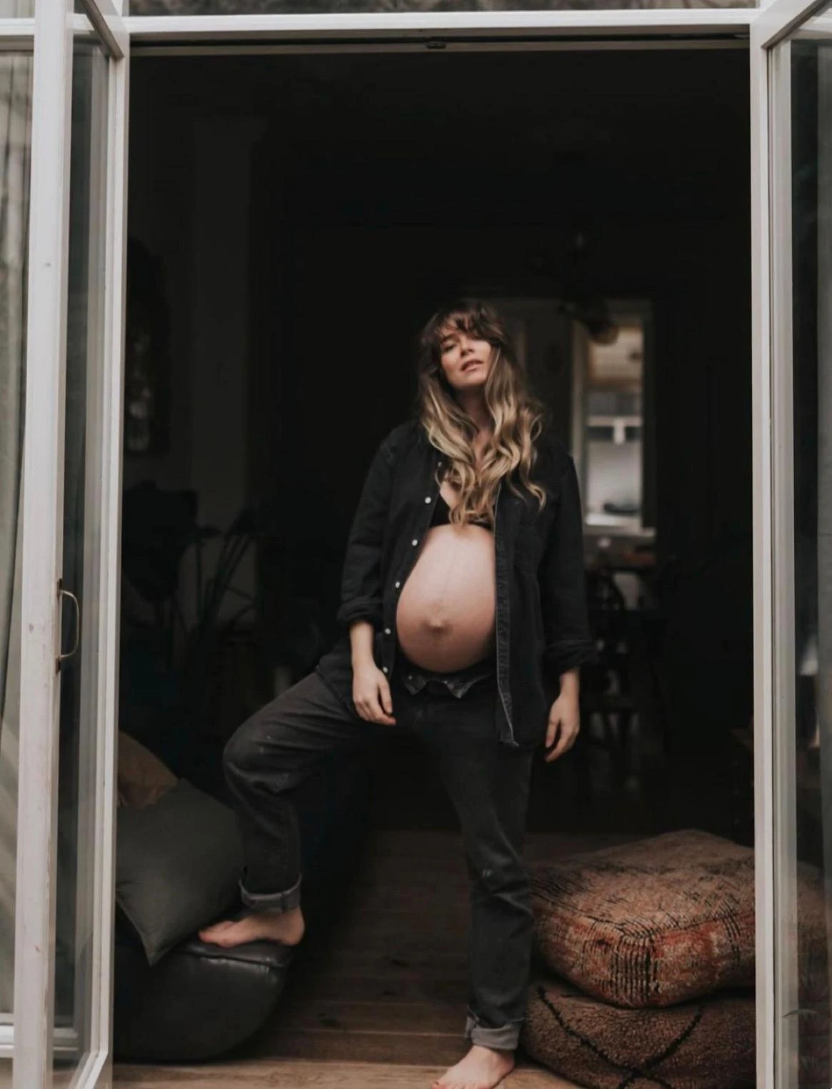 A woman late in pregnancy standing in an open doorway of her home, one foot resting on a floor cushion