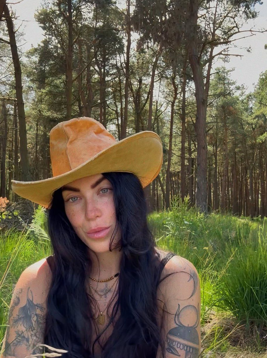 Erin, founder of Feral Kin, sitting in long grass at the edge of a pine wood